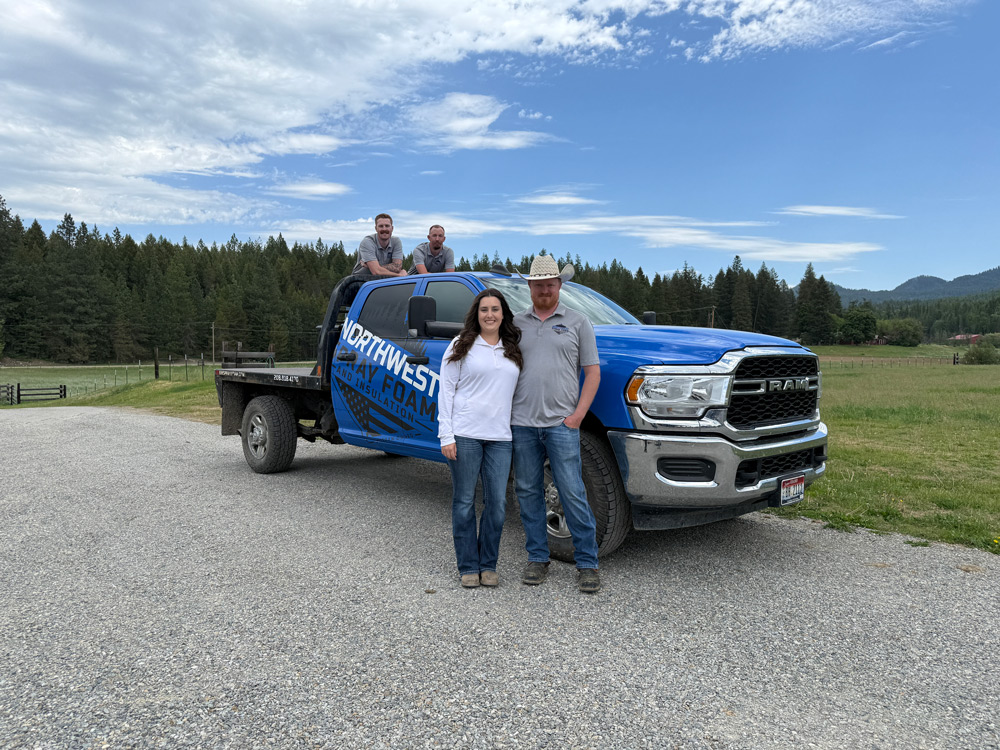 The full crew of four stands with their blue Ram service truck, which features the "Northwest Spray Foam" logo. Two members are in front of the truck while two others are in the truck bed, set against a scenic backdrop of forests and fields.