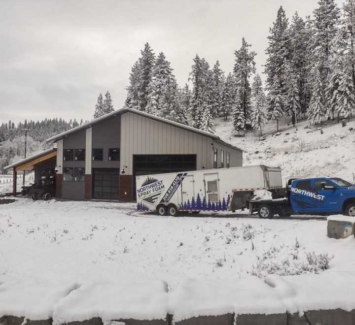 The blue Northwest Spray Foam service truck and branded trailer are parked in the snow outside a modern barndominium-style building. The surrounding landscape, including the pine trees on the hillside, is covered in a fresh layer of snow.