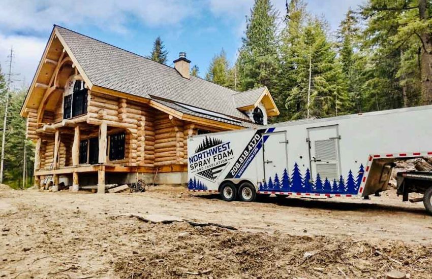 The company's branded trailer is parked on a dirt construction site in front of a large, impressive log home. The scene showcases the company's capability to handle high-end, custom residential projects in rustic settings.